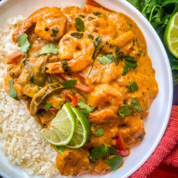 A bowl of creamy Thai shrimp coconut curry with sliced bell peppers, garnished with cilantro and lime wedges, served over white rice. A bunch of fresh cilantro and a lime half are on the side, placed on a dark wooden table. A red cloth is visible nearby.