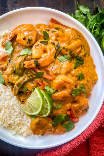 A bowl of creamy Thai shrimp coconut curry with sliced bell peppers, garnished with cilantro and lime wedges, served over white rice. A bunch of fresh cilantro and a lime half are on the side, placed on a dark wooden table. A red cloth is visible nearby.