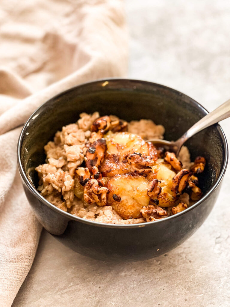 A bowl of banana bread oatmeal is topped with caramelized bananas and roasted cashews, resting on a light textured surface. A spoon sits invitingly in the bowl, while a beige fabric drapes gracefully on the side.