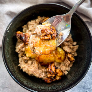 A dark bowl filled with gluten-free oatmeal topped with caramelized bananas and nuts. A spoon lifts a portion from the bowl, reminiscent of banana bread oatmeal. A soft, light-colored cloth is partially visible in the background.