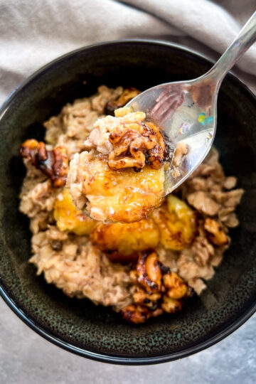 A dark bowl filled with gluten-free oatmeal topped with caramelized bananas and nuts. A spoon lifts a portion from the bowl, reminiscent of banana bread oatmeal. A soft, light-colored cloth is partially visible in the background.