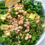 A bowl of Erewhon kale white bean salad, adorned with avocado cubes, grated cheese, and a lemon wedge on the side, sits elegantly on a marble surface.