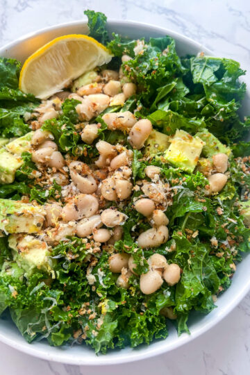 A bowl of Erewhon kale white bean salad, adorned with avocado cubes, grated cheese, and a lemon wedge on the side, sits elegantly on a marble surface.