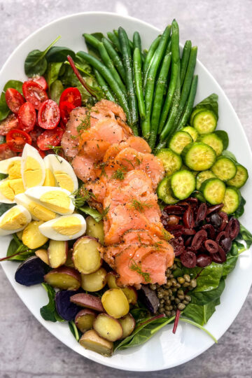 A vibrant oval platter showcases a French Niçoise salad with sliced smoked salmon, cherry tomatoes, hard-boiled eggs, green beans, cucumbers, mixed olives, sliced potatoes, capers, and a bed of spinach leaves arranged artfully.