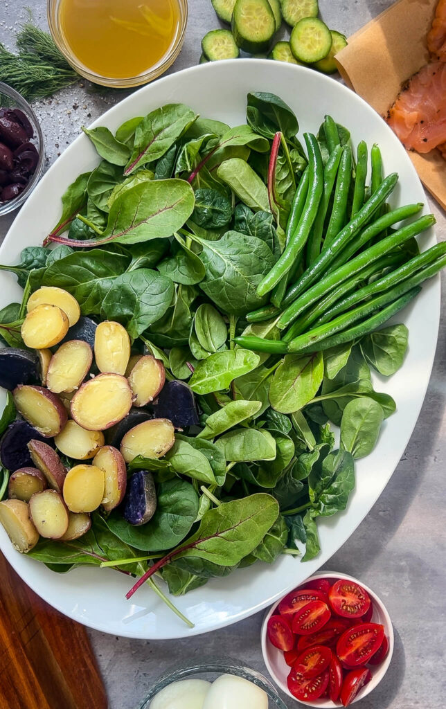 A white oval plate filled with fresh spinach, green beans, and boiled potatoes. Surrounding the plate are bowls of cherry tomatoes, boiled eggs, cucumbers, olives, a small bowl of dressing, and sliced smoked salmon.