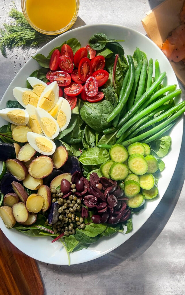 A platter with sections of green beans, sliced cucumbers, halved cherry tomatoes, hard-boiled eggs, roasted potatoes, olives, capers, and fresh spinach leaves, all neatly arranged. A glass of orange juice is in the background.