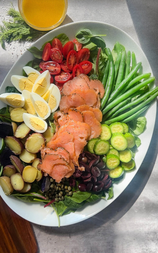 A platter of fresh ingredients: sliced smoked salmon, halved boiled eggs, cherry tomatoes, green beans, sliced cucumbers, small potatoes, dark olives, and capers, arranged on a bed of leafy greens. A small bowl of dressing is beside the platter.