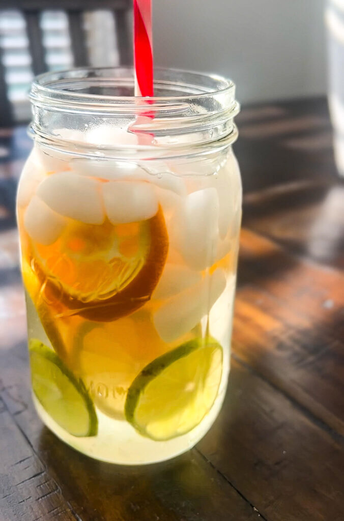 A mason jar filled with iced tea, containing ice cubes and slices of lime and lemon. A red straw is inserted into the jar. The jar is placed on a wooden table with sunlight streaming through in the background.