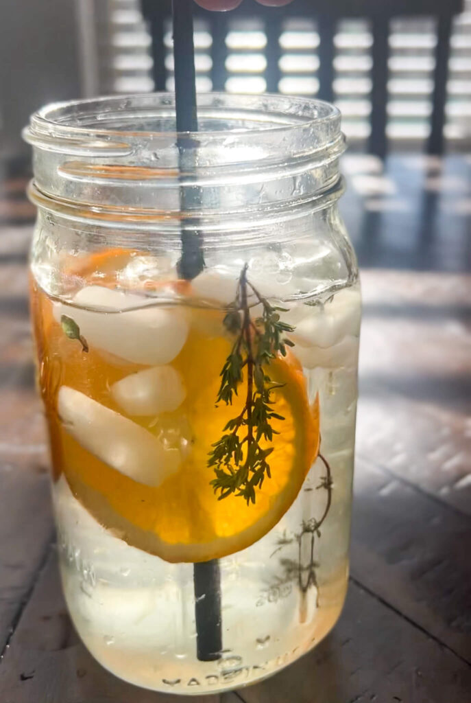 A mason jar filled with a refreshing drink containing ice, a slice of orange, and a sprig of greenery. A black straw stands upright in the jar. Sunlight streams through blinds in the background, casting shadows on the table.