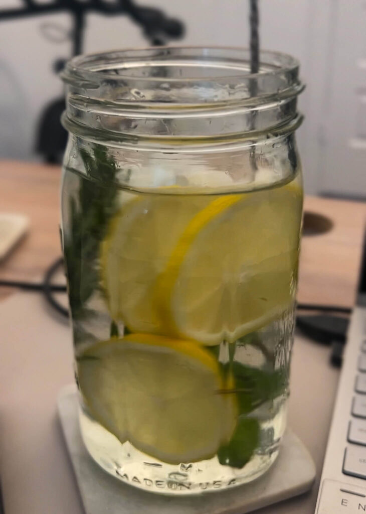 A mason jar filled with water containing lemon and cucumber slices, placed on a coaster near a keyboard. In the background, a blurred view of a bicycle.