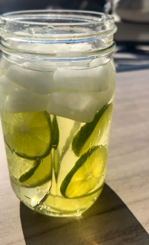 A glass jar filled with lime-infused water and ice cubes sits on a sunlit table. The lime slices are visible through the clear water, and the bright sunlight casts a shadow on the surface.