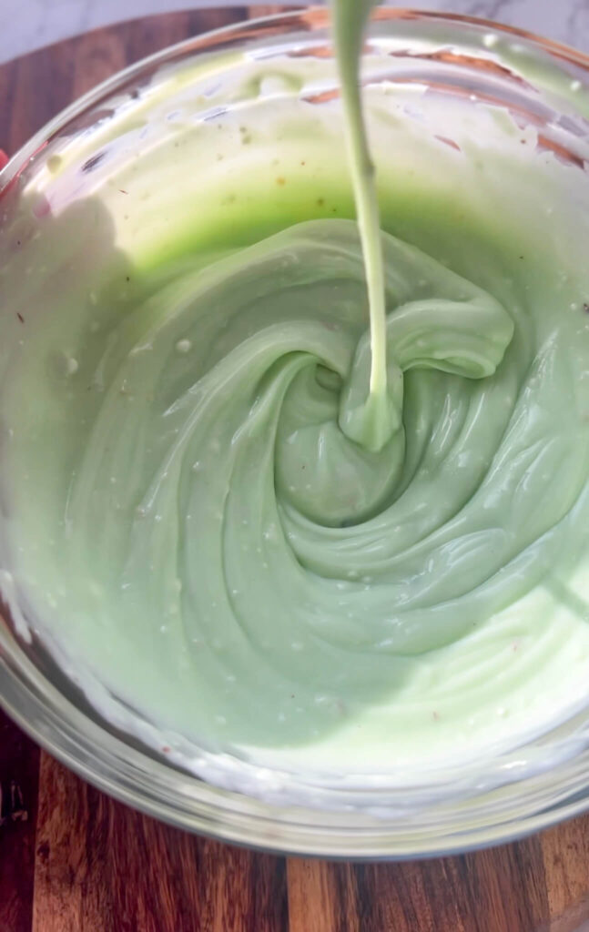 A spoon drizzles creamy, smooth avocado dressing into a clear glass bowl on a wooden surface. The light green mixture swirls, reminiscent of the rich texture found in pistachio cream cheese stuffed dates.