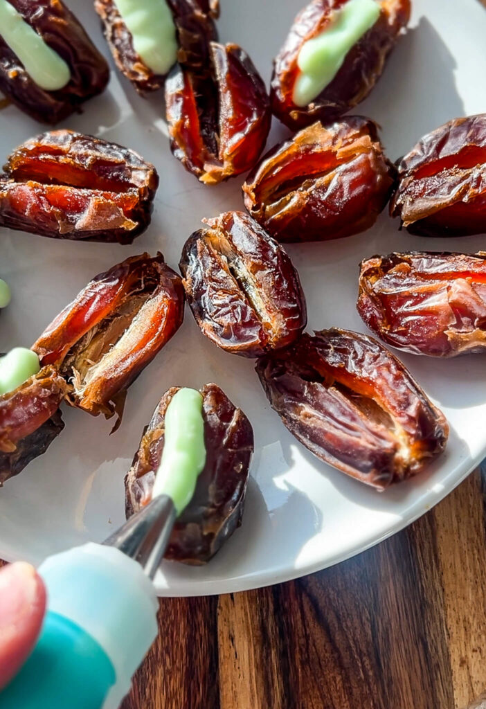 Close-up of a white plate with dates being filled with luscious pistachio cream cheese from a piping bag. These delectable dates, opened and ready, bask in the warm sunlight casting an inviting glow on the wooden table—perfect for your next stuffed dates recipe.