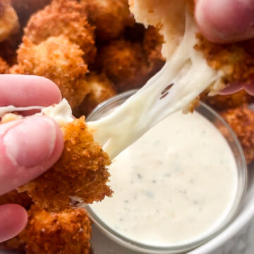 Close-up of hands pulling apart fried, breaded mozzarella sticks, showing melted cheese stretching between them. In the background, a bowl of Nashville hot mozzarella sticks and a small bowl of dipping sauce are visible.