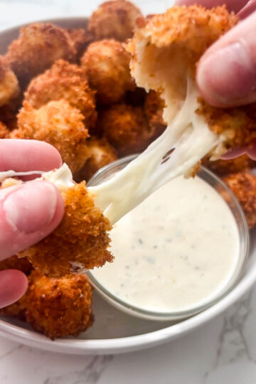 Close-up of hands pulling apart fried, breaded mozzarella sticks, showing melted cheese stretching between them. In the background, a bowl of Nashville hot mozzarella sticks and a small bowl of dipping sauce are visible.