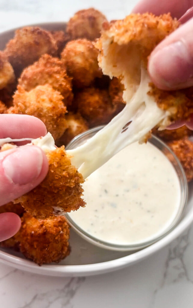 Close-up of hands pulling apart fried, breaded mozzarella sticks, showing melted cheese stretching between them. In the background, a bowl of Nashville hot mozzarella sticks and a small bowl of dipping sauce are visible.