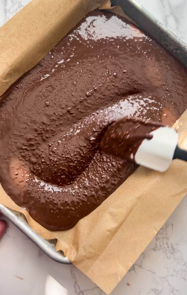 A spatula spreads thick, glossy chocolate batter for quinoa chocolate crunch bars in a parchment-lined square baking pan on a marble countertop.