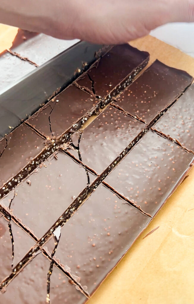 Close-up of a hand cutting a large slab of chocolate quinoa crunch bar into smaller rectangular pieces on a sheet of parchment paper. The chocolate appears dark with a glossy texture and some visible speckles or texture within.