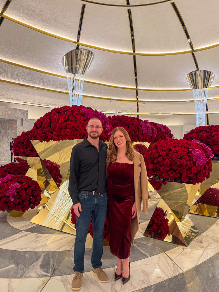 A man and woman standing in front of large, decorative red rose arrangements with mirrored accents. The woman is wearing a burgundy dress and beige coat, and the man is in a black shirt and jeans. The ceiling features modern light fixtures.