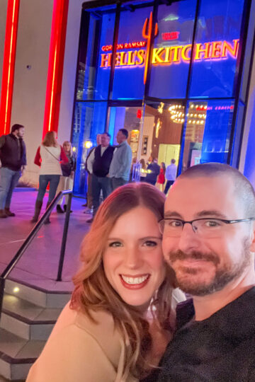 A couple smiling and taking a selfie outside a restaurant entrance lit by red and blue neon lights with Gordon Ramsay Hells Kitchen signage above. Several people are gathered around the entrance in the background.