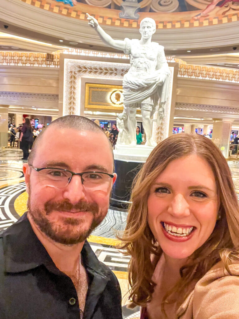 A man and woman smile for a selfie in an ornate indoor setting with a large statue of a robed figure in the background. The statue has a raised arm and stands on a decorative pedestal under a domed ceiling.