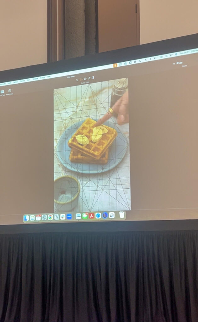 A large screen displays an image of a person pouring syrup over a stack of waffles topped with butter. The screen shows a desktop interface with various icons. The setup is in a room with a black tablecloth-covered table below the screen.