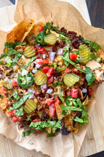 A loaded nacho platter on brown parchment paper. Topped with cheese, sliced pickles, jalapeños, shredded lettuce, diced tomatoes, chopped red onions, and bits of bacon. Placed on a wooden board.