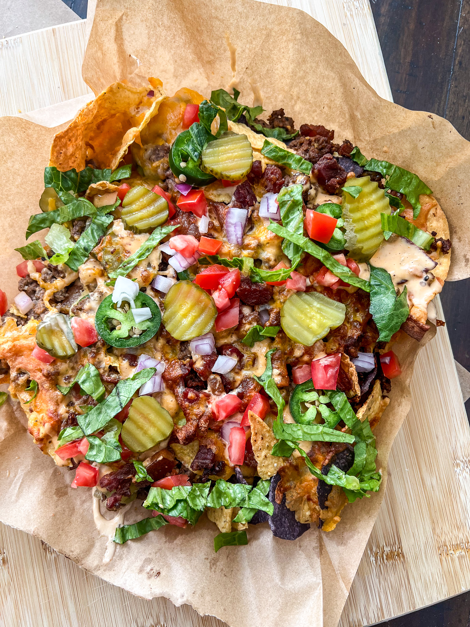 A pizza topped with pickles, jalapeños, diced tomatoes, red onions, lettuce, and crispy bits, on a piece of parchment paper. The pizza has a variety of toppings including both fresh vegetables and savory elements.