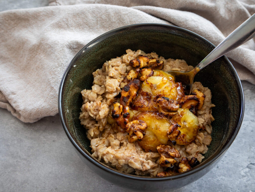 A bowl of gluten-free banana bread oatmeal topped with caramelized bananas and walnuts. A spoon rests inside the bowl, and a beige cloth napkin is placed underneath it on a gray surface.