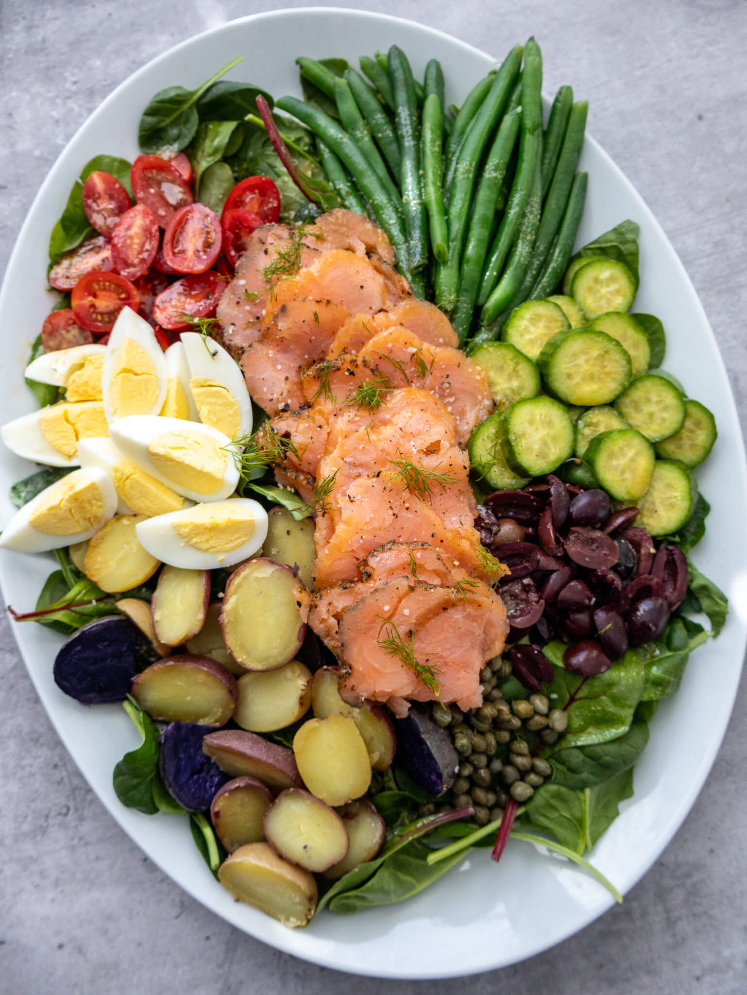 An oval platter with smoked salmon, cherry tomatoes, green beans, cucumber slices, hard-boiled eggs, olives, capers, and assorted potatoes on a bed of leafy greens.