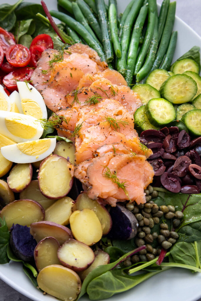 A vibrant salad with smoked salmon, green beans, sliced cucumbers, cherry tomatoes, hard-boiled eggs, olives, potatoes, capers, and spinach, all garnished with fresh dill.