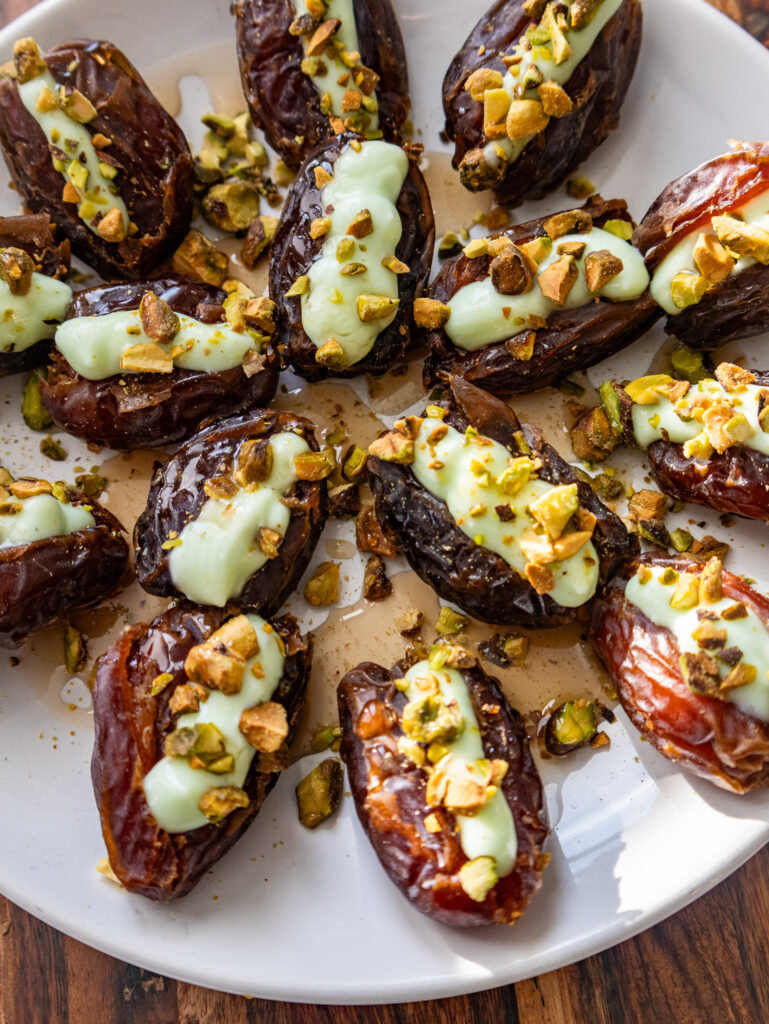 A delightful plate of pistachio cream stuffed dates, topped with a velvety green cream and sprinkled with chopped pistachios. This elegant dish is beautifully arranged on a white plate against the natural charm of a wooden table surface.