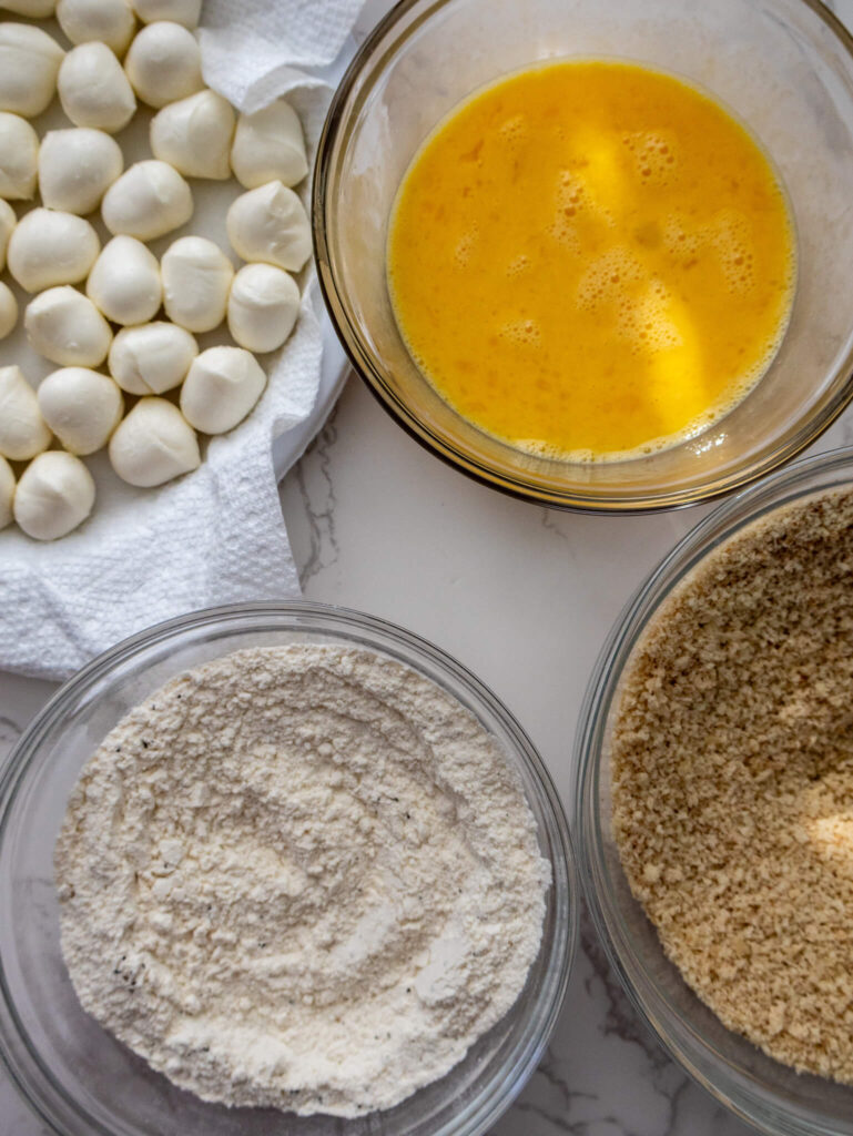 A dredging station with mozzarella balls in 1 bowl, whisked eggs in another, seasoned panko breadcrumbs in another, and flour in the other bowl. 