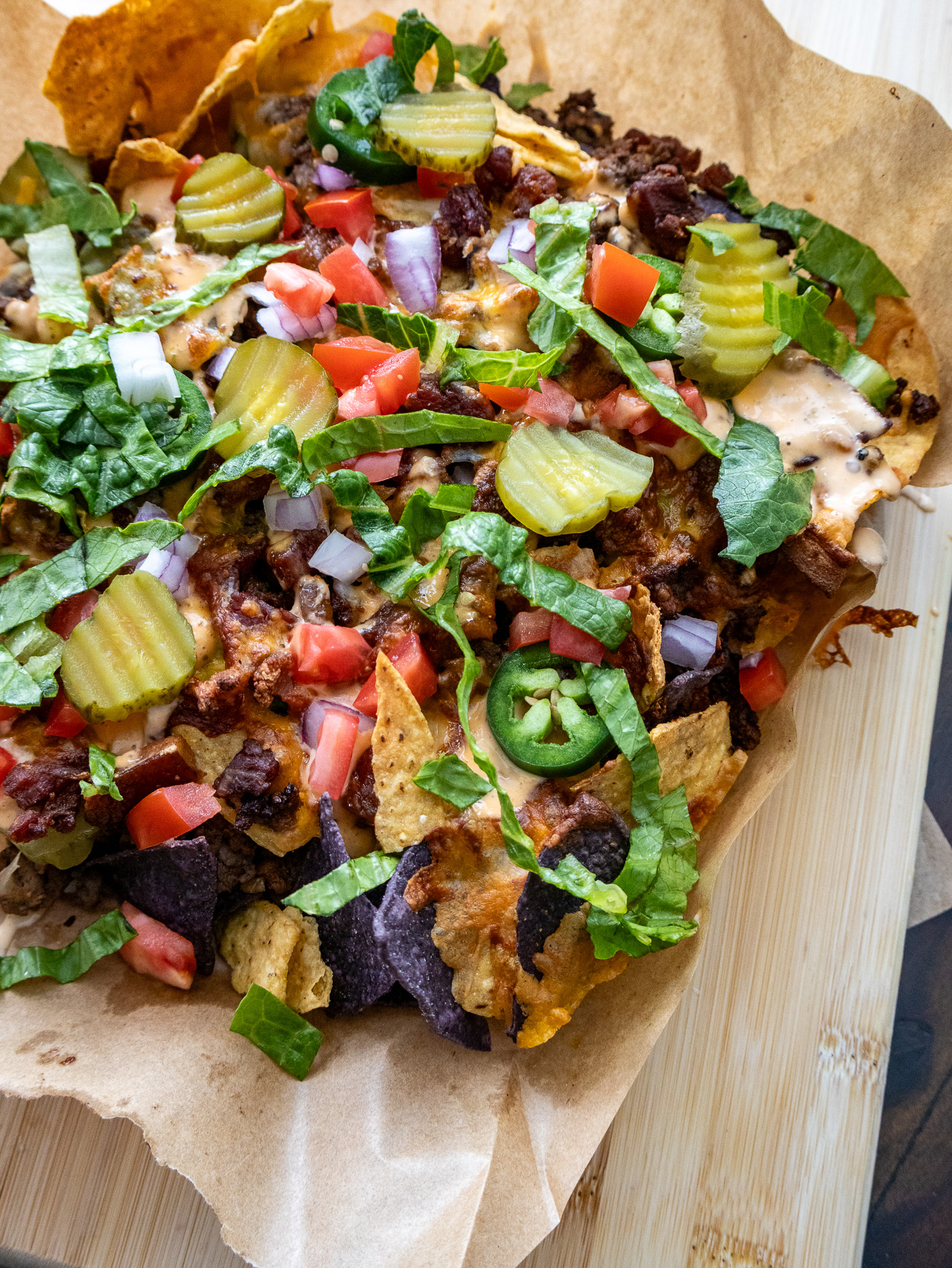 A tray of loaded nachos topped with sliced pickles, tomatoes, lettuce, onions, jalapeños, and drizzled with a creamy sauce. The nachos rest on parchment paper over a wooden surface.