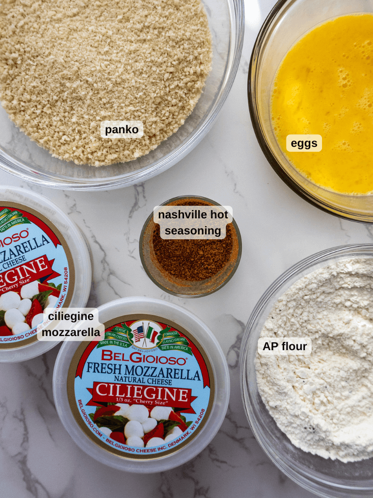 Ingredients for mozzarella bites include panko breadcrumbs, beaten eggs, Nashville hot seasoning, AP flour, and fresh ciliegine mozzarella. All items are arranged in bowls and containers on a marble surface, perfect for crafting your own Nashville hot mozzarella sticks.