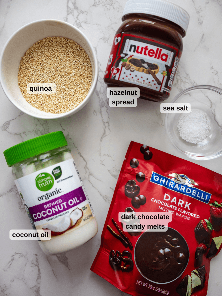 Top-down view of ingredients on a marble surface: a bowl of quinoa for delicious quinoa crunch bars, jar of hazelnut spread, small dish of sea salt, jar of organic coconut oil, and a bag of dark chocolate candy melts.