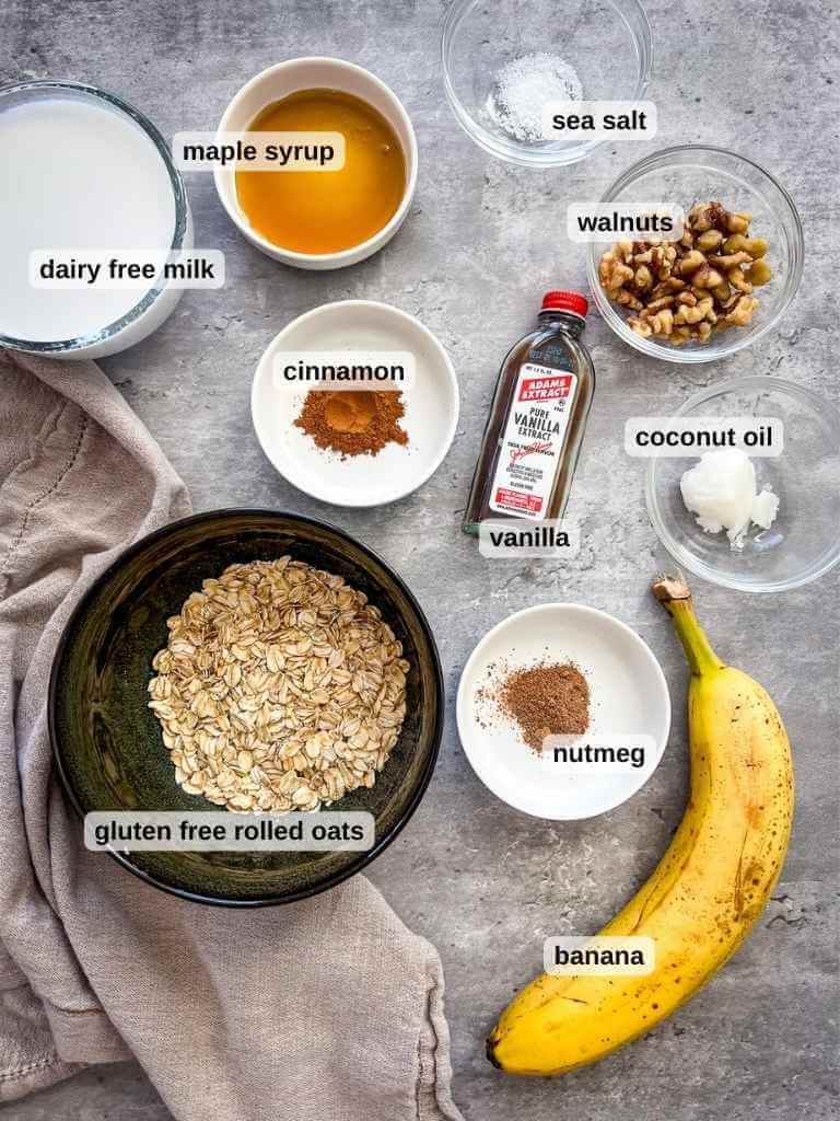 Top-down view of ingredients for banana bread oatmeal on a gray surface: dairy-free milk, maple syrup, walnuts, sea salt, coconut oil, gluten-free rolled oats, cinnamon, nutmeg, vanilla extract, and a banana. A textured cloth lies to the side.