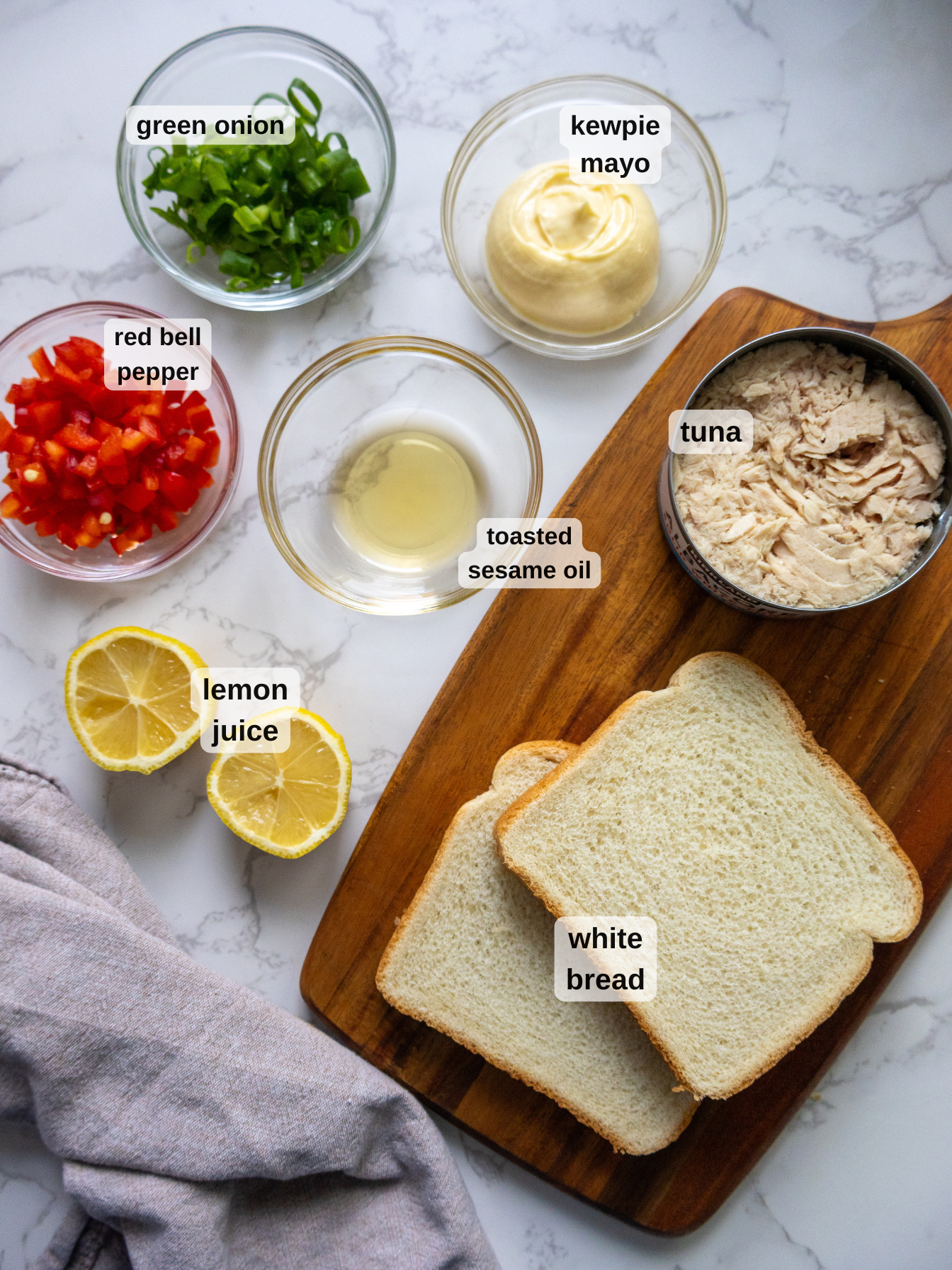 Ingredients on a marble countertop include sliced green onions, Kewpie mayo, red bell peppers, sesame oil, lemon halves, lemon juice, tuna in a tin, and white bread slices on a wooden board. A gray cloth napkin is on the side.