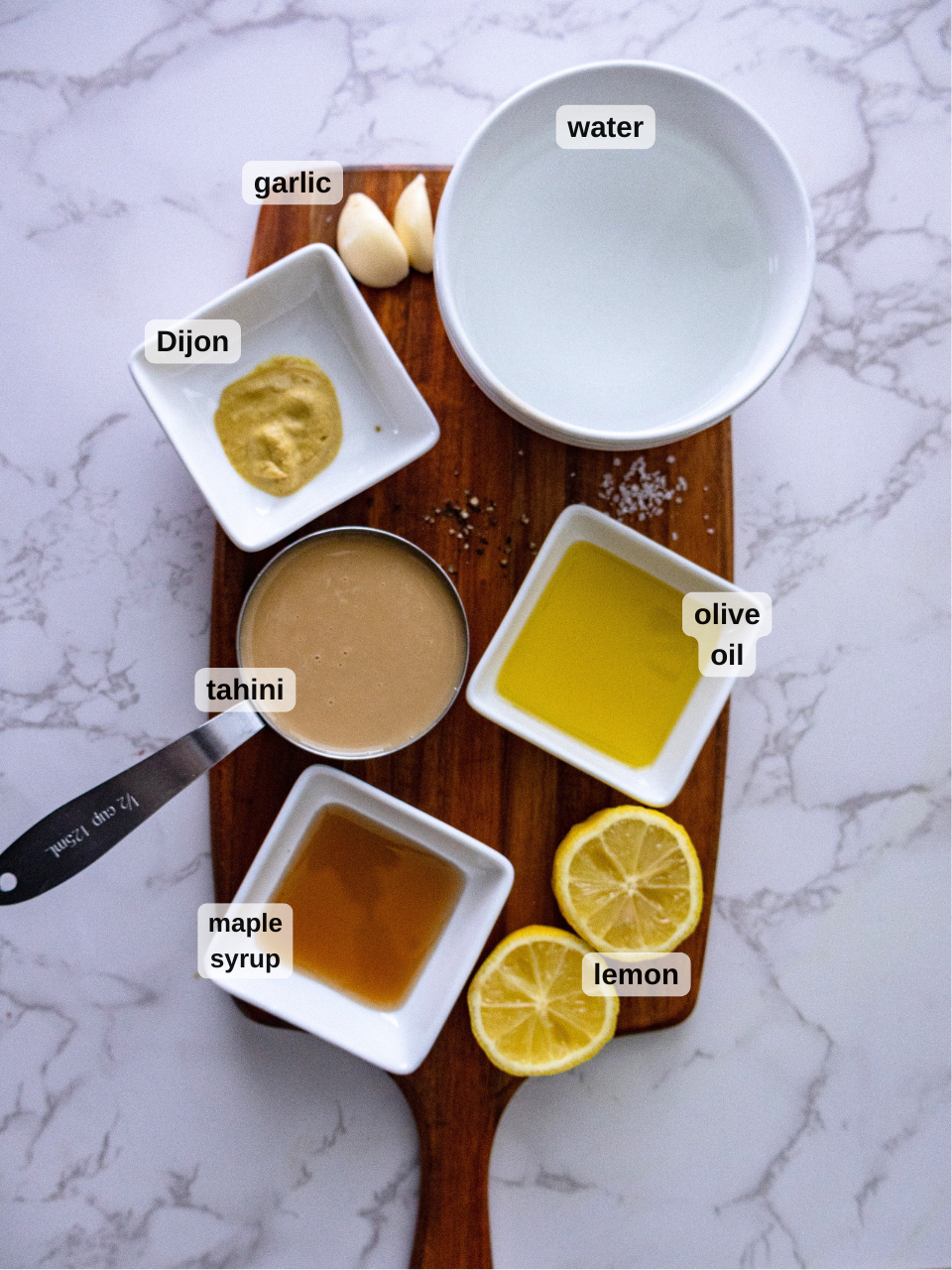 A wooden board on a marble surface showcases ingredients for a crunchy quinoa salad: a bowl of water, garlic cloves, Dijon mustard, tahini dressing, olive oil, maple syrup, lemon slices, and salt. Nearby lies a knife.