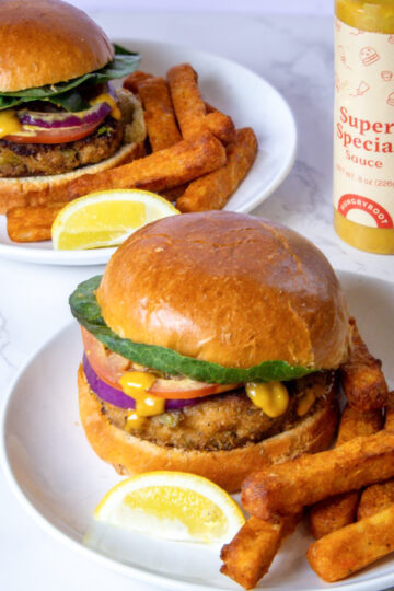 Two plates with veggie burgers topped with lettuce, tomato, onion, and mustard. Each plate has thick-cut fries and a lemon wedge on the side. A bottle of Super Special sauce sits in the background on a marble surface next to a crab cake sandwich.