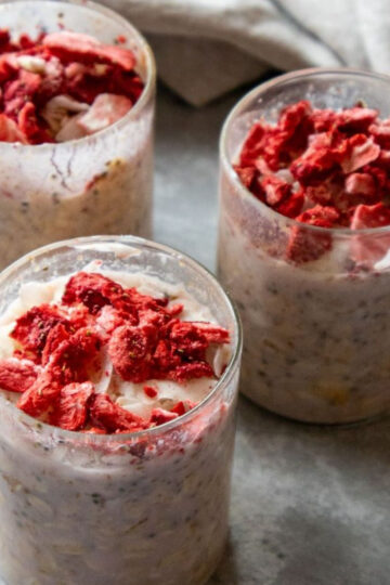 Three glasses filled with creamy strawberry milk overnight oats are topped with chunks of red fruit, likely strawberries. The glasses are placed on a light gray surface, with a soft focus background.