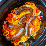 Five cooked sausages sit in a slow cooker surrounded by sliced red, orange, and yellow bell peppers. The dish is garnished with cooked onions, creating a colorful and appetizing presentation.