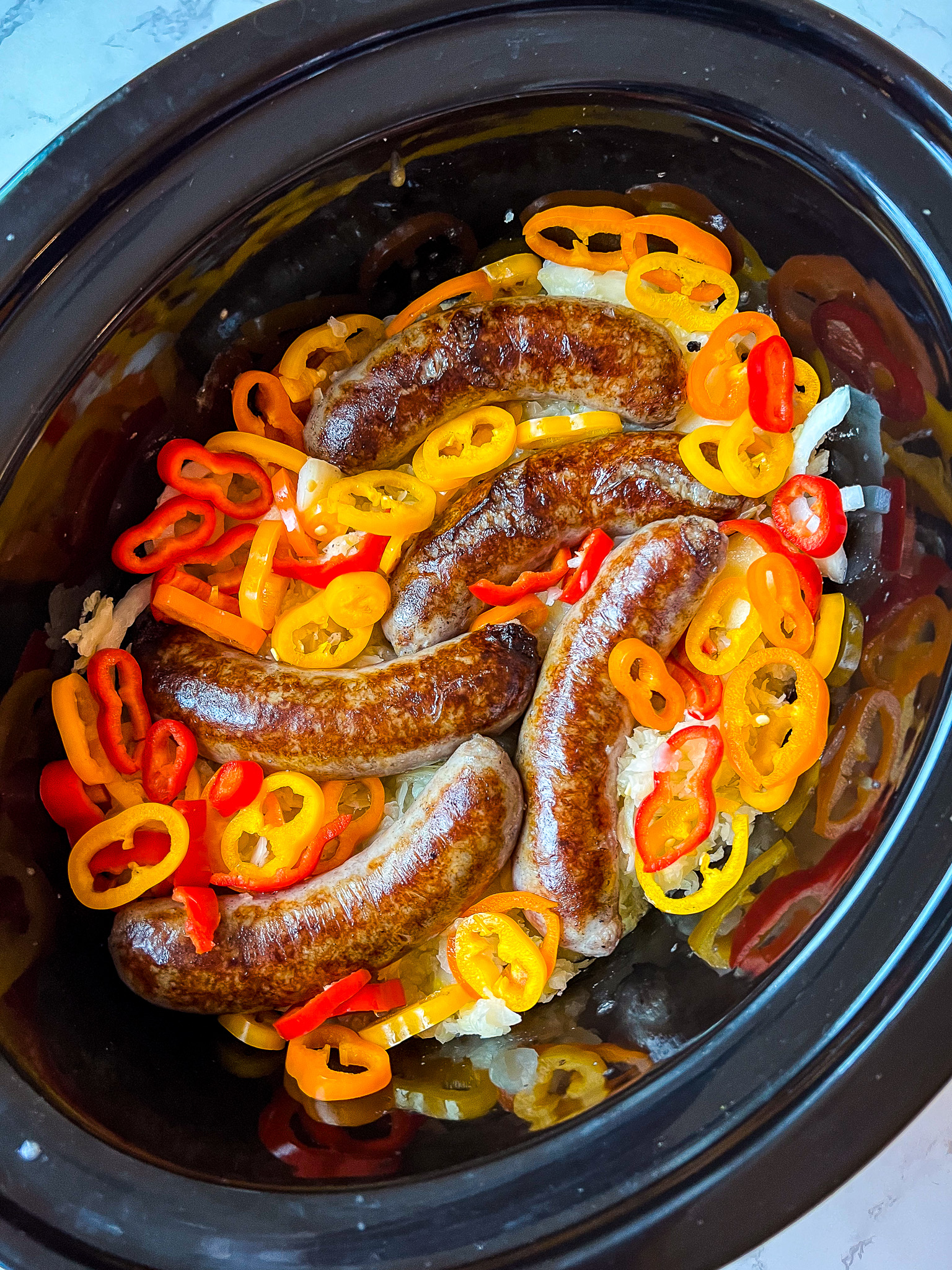 Five cooked Beer Brats sit in a slow cooker surrounded by sliced red, orange, and yellow bell peppers. The dish is garnished with cooked onions, creating a colorful and appetizing presentation.