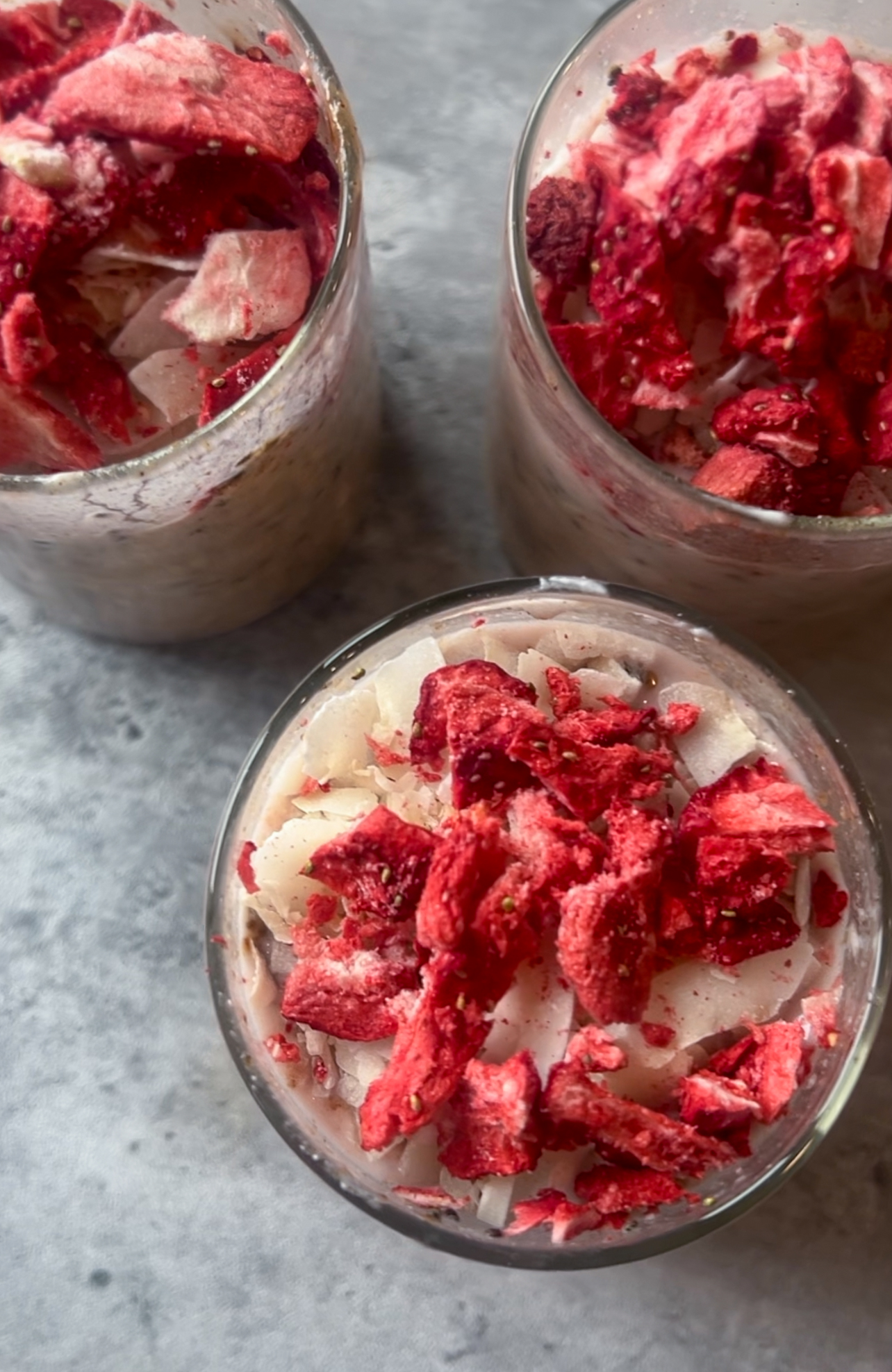 Three glasses filled with strawberry milk overnight oats topped with dried strawberries and coconut flakes are arranged on a textured gray surface. The vibrant red of the strawberries contrasts beautifully with the creamy oats and white coconut.