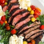 Sliced flank steak cooked to medium-rare with a rich coffee rub, served on a white plate alongside roasted cherry tomatoes, fresh arugula, and dollops of creamy burrata cheese.