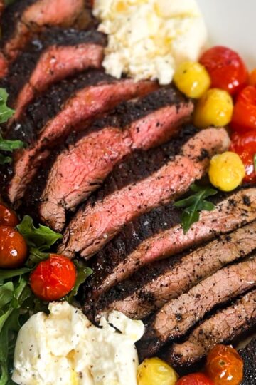 Sliced flank steak cooked to medium-rare with a rich coffee rub, served on a white plate alongside roasted cherry tomatoes, fresh arugula, and dollops of creamy burrata cheese.