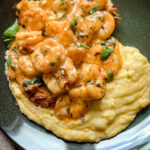 A plate of creamy parmesan polenta topped with seasoned shrimp and garnished with fresh herbs, served on a dark dish. Perfect for a Valentines Day dinner, the shrimp cooked in a rich, spicy sauce add contrast to the smooth texture of the polenta.