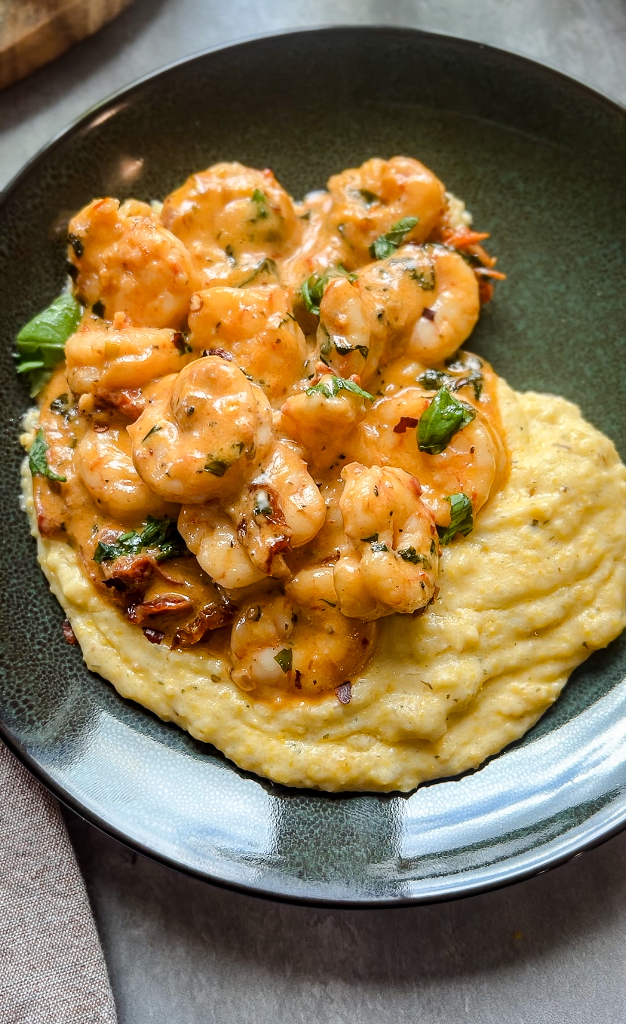 A plate of creamy parmesan polenta topped with seasoned shrimp and garnished with fresh herbs, served on a dark dish. Perfect for a Valentines Day dinner, the shrimp cooked in a rich, spicy sauce add contrast to the smooth texture of the polenta.