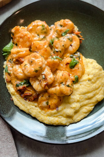 A black plate filled with creamy Parmesan grits topped with seasoned shrimp and garnished with chopped herbs, reminiscent of an Easy Marry Me Shrimp dish. This savory presentation is perfect for a special Valentines Day dinner.