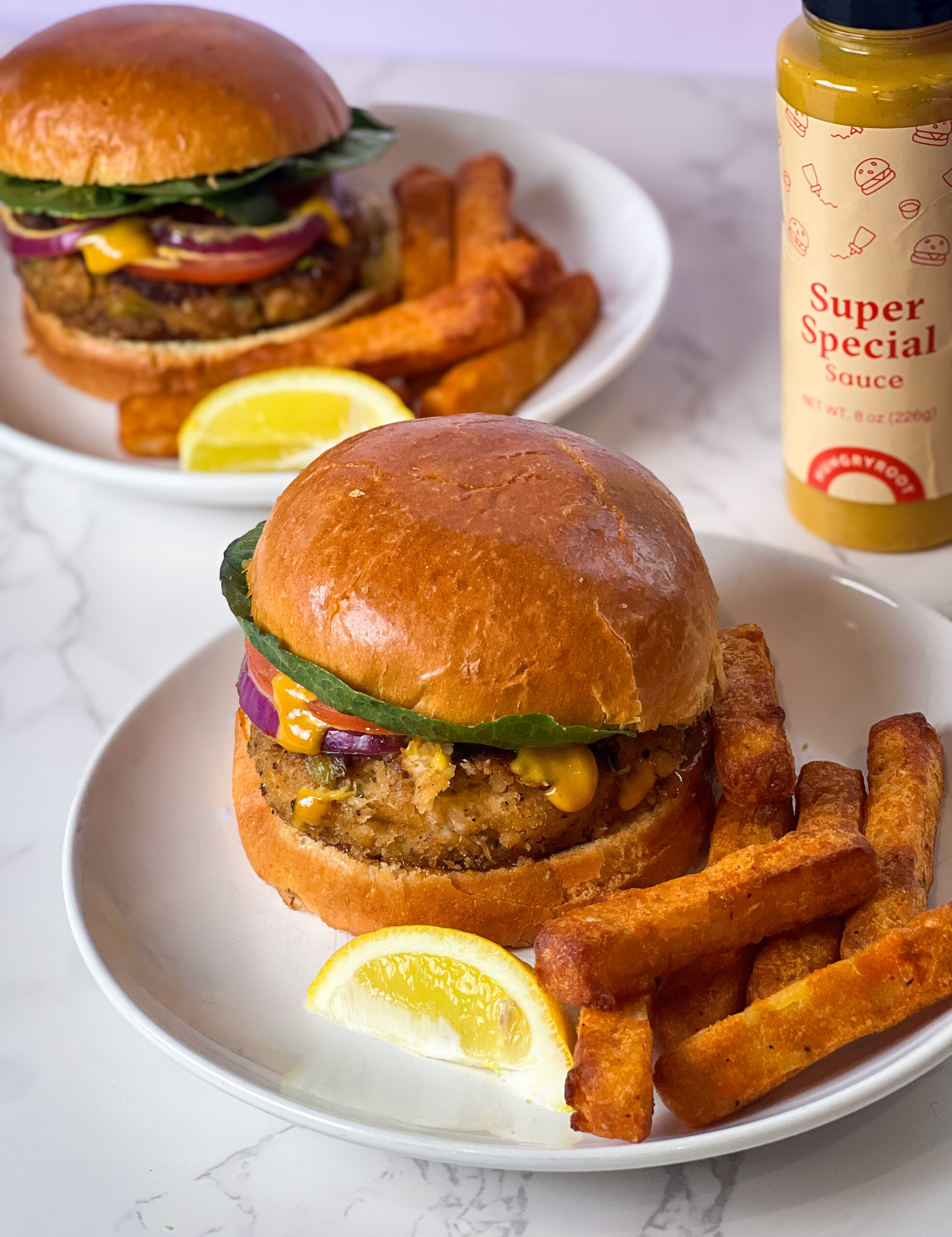 Two plates of chicken sandwiches with lettuce, tomato, and sauce sit next to thick-cut fries and lemon wedges. A bottle of Super Special Sauce is nearby on a white surface.